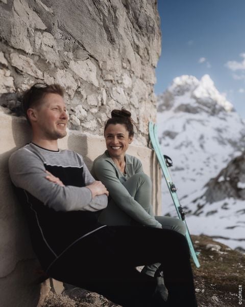 Zwei Personen in Winterkleidung sitzen hinten in einem auf einer verschneiten Straße geparkten Fahrzeug. Beide lächeln. Die Person links trägt eine schwarze Mütze und ein Sweatshirt, während die Person rechts einen gemusterten Pullover, einen Helm und eine Schutzbrille trägt. Text auf dem Bild.