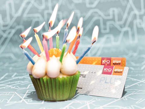 Ein grüner Cupcake mit weißem Zuckerguss und brennenden Kerzen steht vor zwei Kreditkarten. Der Hintergrund zeigt ein hellblaues abstraktes Muster.