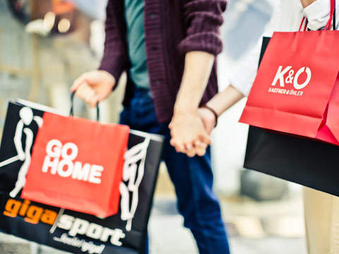 Zwei Personen halten sich an den Händen und tragen Einkaufstüten. Auf den Tüten sind verschiedene Logos zu sehen, darunter „GO HOME“ und „K & Ö“. Sie gehen im Freien spazieren und die Szene ist hell beleuchtet, was ein lebhaftes Einkaufserlebnis suggeriert.