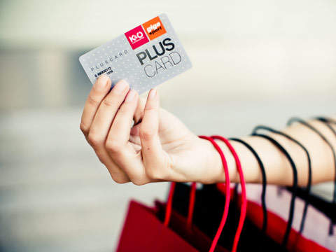 Eine Hand hält eine PLUSCARD mit sichtbaren KID- und Click-Logos. Am Arm befinden sich mehrere Einkaufstüten mit roten und schwarzen Griffen, die eine Einkaufsszene suggerieren. Der Hintergrund ist unscharf.