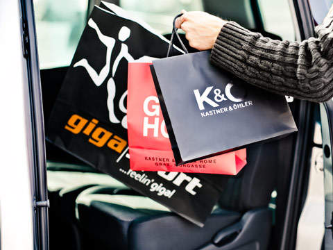 Eine Person hält Einkaufstüten vor einer offenen Autotür. Die Tüten sind schwarz und rot und mit Logos und Texten versehen, die auf kürzlich getätigte Einkäufe hinweisen. Eine Person trägt einen dunklen Pullover und der Innenraum des Autos ist sichtbar.