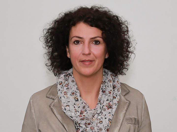 Eine Person mit lockigem dunklem Haar, die einen geblümten Schal und einen beigen Blazer trägt, blickt direkt in die Kamera. Der Hintergrund ist schlicht und neutral.