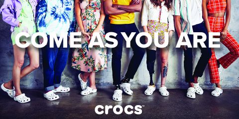 Eine bunt gemischte Gruppe von Menschen steht an einer Betonwand. Jeder trägt ein anderes Outfit und weiße Crocs. Der Text „COME AS YOU ARE“ ist deutlich über dem Crocs-Logo zu sehen.