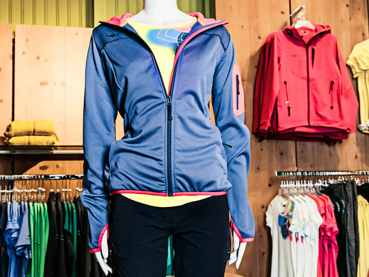 Eine Schaufensterpuppe trägt eine hellblaue Jacke mit rosa Reißverschluss und Besatz über einem gelben Hemd und einer schwarzen Hose. Dahinter befinden sich Holzregale und -ständer, auf denen Jacken und Hemden in verschiedenen Farben in einem Laden ausgestellt sind.