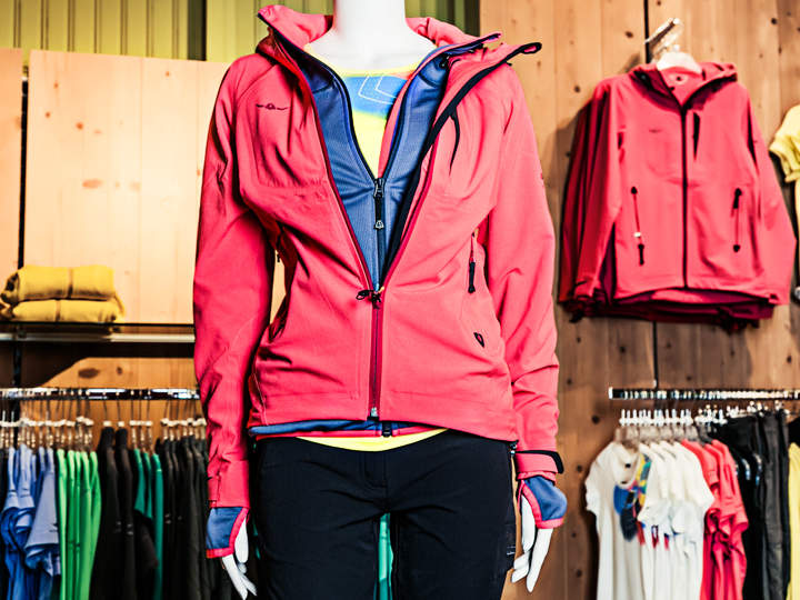 Eine Schaufensterpuppe präsentiert ein mehrlagiges Outfit mit einer rosa Jacke über einer blauen und bunten Unterhemden. Sie trägt schwarze Hosen und ist von ähnlicher Kleidung auf Ständern in einem Geschäft umgeben. Im Hintergrund sind Holzpaneele zu sehen.