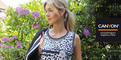 Eine Frau in einem ärmellosen Oberteil mit schwarz-weißem Muster steht im Freien neben blühenden lila Blumen und Grünpflanzen. Auf der rechten Seite ist das Logo „CANYON women sports“ zu sehen. Sie hält eine schwarze Jacke über der Schulter.