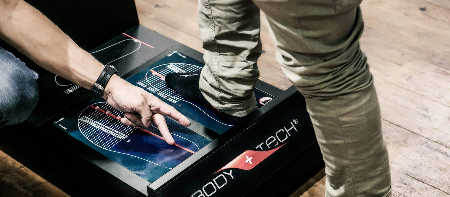 Eine Person steht auf einem Fußscanner, während eine andere Person auf die digitale Anzeige zeigt, die die Fußdruckanalyse anzeigt. Das Gerät trägt die Aufschrift „Body Tech“ und steht auf einem Holzboden.