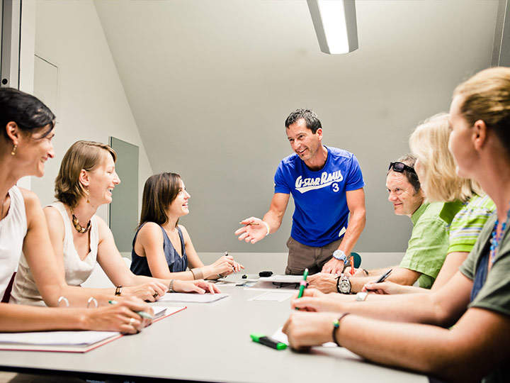 Eine Gruppe von sechs Personen sitzt mit Notizbüchern, Stiften und aufgeklappten Laptops um einen Tisch. Eine Person in einem blauen T-Shirt steht da, lächelt und gestikuliert, als würde sie zur Gruppe sprechen. Es scheint sich um eine Besprechung oder Diskussion zu handeln.