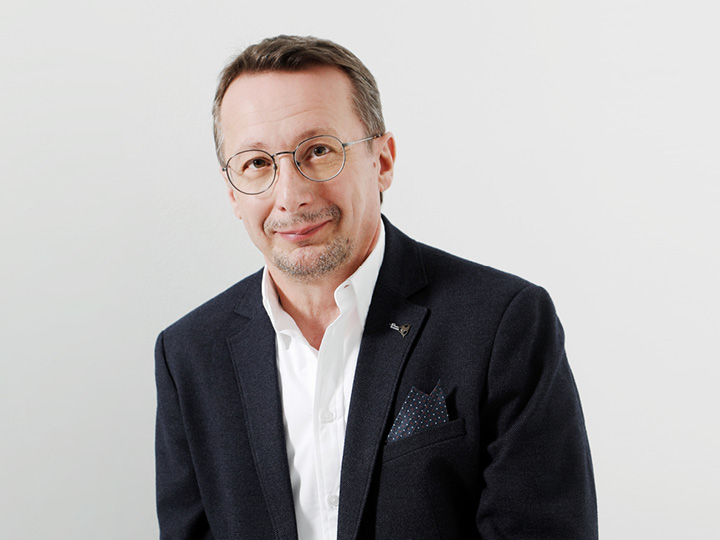 Mann mit kurzen braunen Haaren und Brille, trägt einen dunklen Blazer und ein weißes Hemd, lächelt vor einem schlichten Hintergrund.