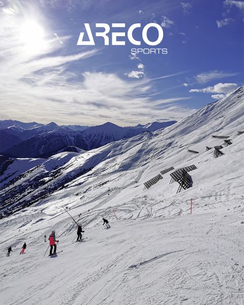 Skifahrer auf einem schneebedeckten Berghang unter blauem Himmel mit vereinzelten Wolken. Die Sonne scheint hell und wirft Schatten auf den Schnee. Oben steht der Text „ARECO SPORTS“. Im Hintergrund sind schneebedeckte Gipfel zu sehen.