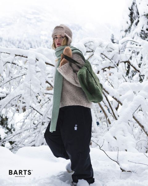 Eine Person mit beigefarbener Strickmütze, Pullover, grünem Schal und schwarzer Hose steht in einer verschneiten Landschaft und trägt einen grünen Rucksack. Schneebedeckte Äste und Bäume prägen den Hintergrund. Der Text auf dem Bild lautet „BARTS AMSTERDAM“.