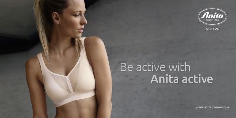 Frau im Sport-BH steht vor grauem Hintergrund und schaut zur Seite. Der Text lautet „Sei aktiv mit Anita Active.“ Das Anita-Logo und der Website-Link befinden sich in der Ecke.
