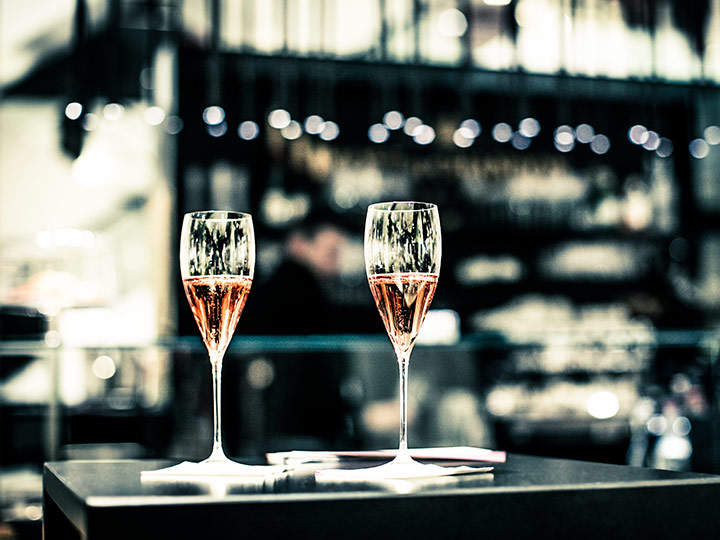 Zwei Gläser Champagner stehen auf einem Tisch in einer schwach beleuchteten Bar. Der Hintergrund ist verschwommen, mit funkelnden Lichtern und Regalen voller Flaschen, was eine gemütliche und intime Atmosphäre schafft.