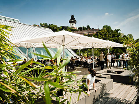 Eine Dachterrasse mit Menschen, die unter großen weißen Sonnenschirmen sitzen und stehen. Im Vordergrund sind grüne Pflanzen zu sehen, und im Hintergrund ist unter einem klaren blauen Himmel ein Uhrturm auf einem Hügel zu sehen.