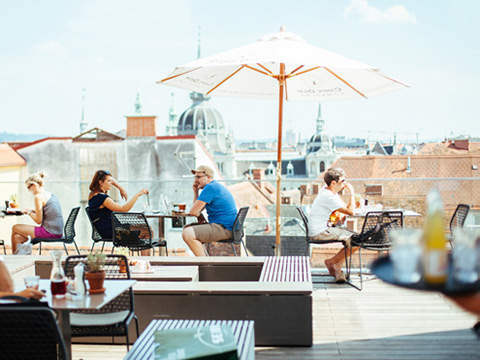 Menschen sitzen und unterhalten sich an Tischen auf einer Dachterrasse unter Sonnenschirmen. Die Szene ist hell und sonnig, mit Blick auf die Dächer der Stadt und entfernte Gebäude im Hintergrund. Im Vordergrund ist teilweise ein Tablett mit Getränken zu sehen.