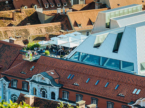 Luftaufnahme einer Stadtlandschaft mit einer Mischung aus traditionellen Gebäuden mit roten Dächern und einer modernen Struktur mit eckigen Fenstern. Zu sehen ist eine Dachterrasse mit weißen Sonnenschirmen und Menschen, umgeben von städtischer Architektur.