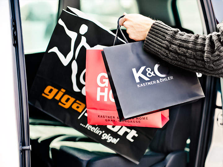 Eine Person hält zwei schwarze Einkaufstüten, eine von K & Ö Kastner & Öhler und eine andere mit einem grafischen Logo, und lädt sie in ein Auto. Der Innenraum des Rücksitzes ist sichtbar.