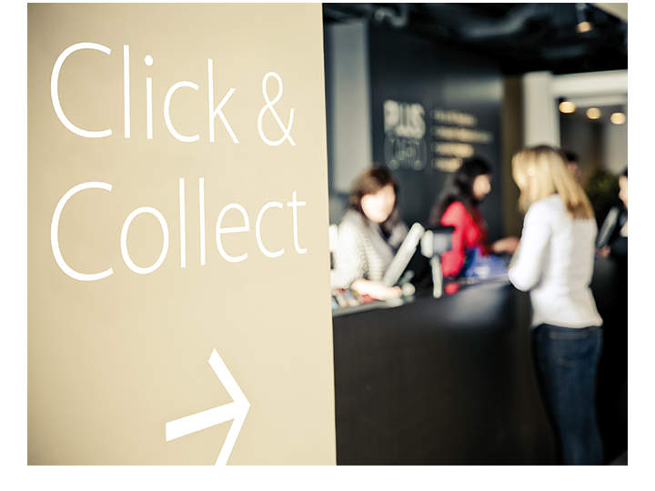 Schild mit der Aufschrift „Click & Collect“ und einem nach rechts zeigenden Pfeil. Im Hintergrund eine Theke mit drei Personen, die miteinander interagieren, wahrscheinlich in einem Laden. Die Szene ist leicht verschwommen, um einen Weichzeichnereffekt zu erzielen.