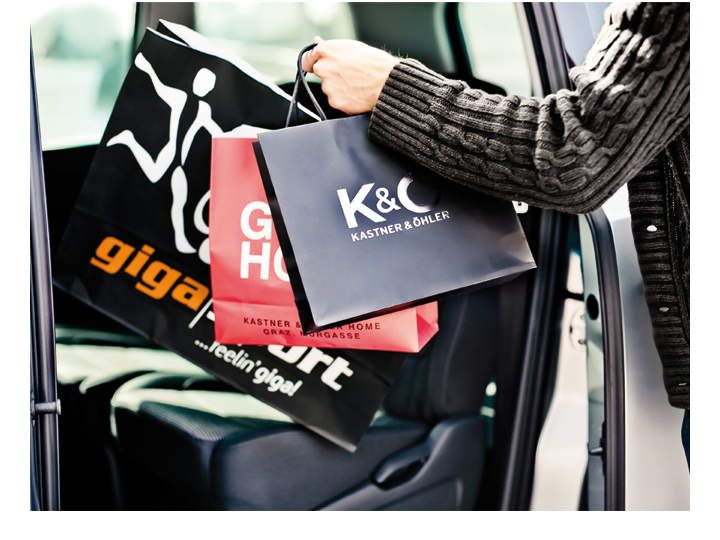 Eine Person hält mehrere Einkaufstüten mit Logos von Kastner & Öhler und anderen Marken und legt sie auf den Rücksitz eines Autos. Die Tüten sind schwarz und rot, der Innenraum des Fahrzeugs ist teilweise sichtbar.
