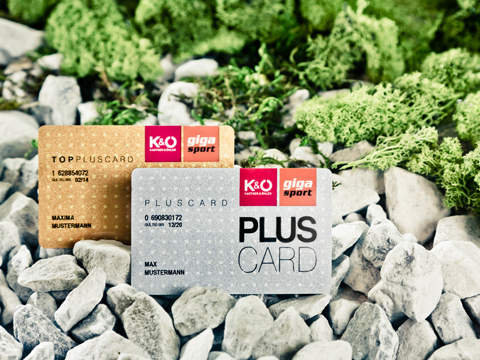 Zwei Karten auf einem Bett aus grauen Steinen mit grünem Moos. Auf den Karten steht „TOPPLUSECARD“ und „PLUSCARD“, dazu ein rotes K&Ö-Logo. Auf beiden Karten ist der Name „MAX MUSTERMANN“ zu sehen. Das Design ist minimalistisch und betont den Premium-Branding-Charakter.