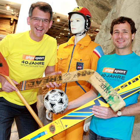 Zwei Männer in gelben und blauen Hemden posieren mit einer lebensgroßen Schaufensterpuppe in orangefarbener Sportkleidung. Sie halten alte Sportgeräte in der Hand, darunter einen Hockeyschläger, Skier und einen Fußball. Die Szene scheint drinnen vor einer felsigen Kulisse zu sein.
