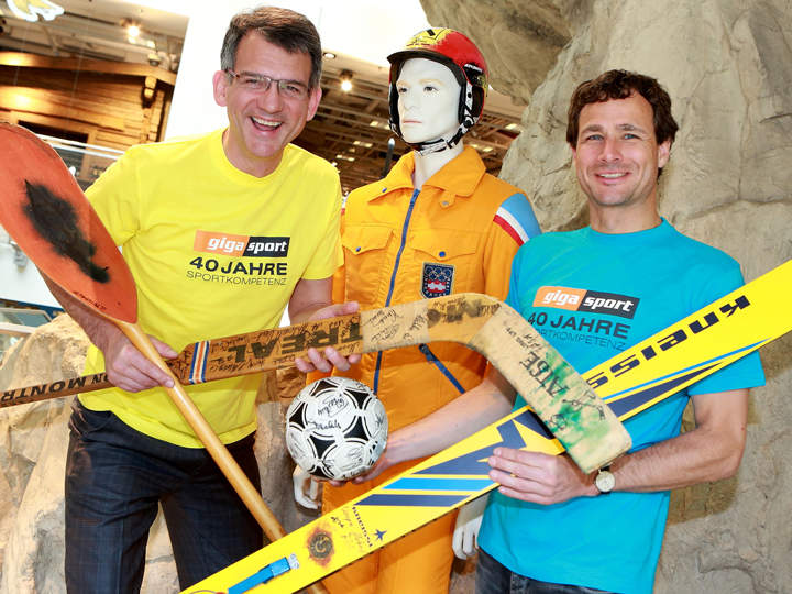 Zwei lächelnde Männer in gelben T-Shirts halten Sportgeräte in der Hand, darunter ein Kajakpaddel, Skier und einen Hockeyschläger. Sie stehen neben einer Schaufensterpuppe in orangefarbenem Skianzug und Helm vor einer felsenähnlichen Kulisse.