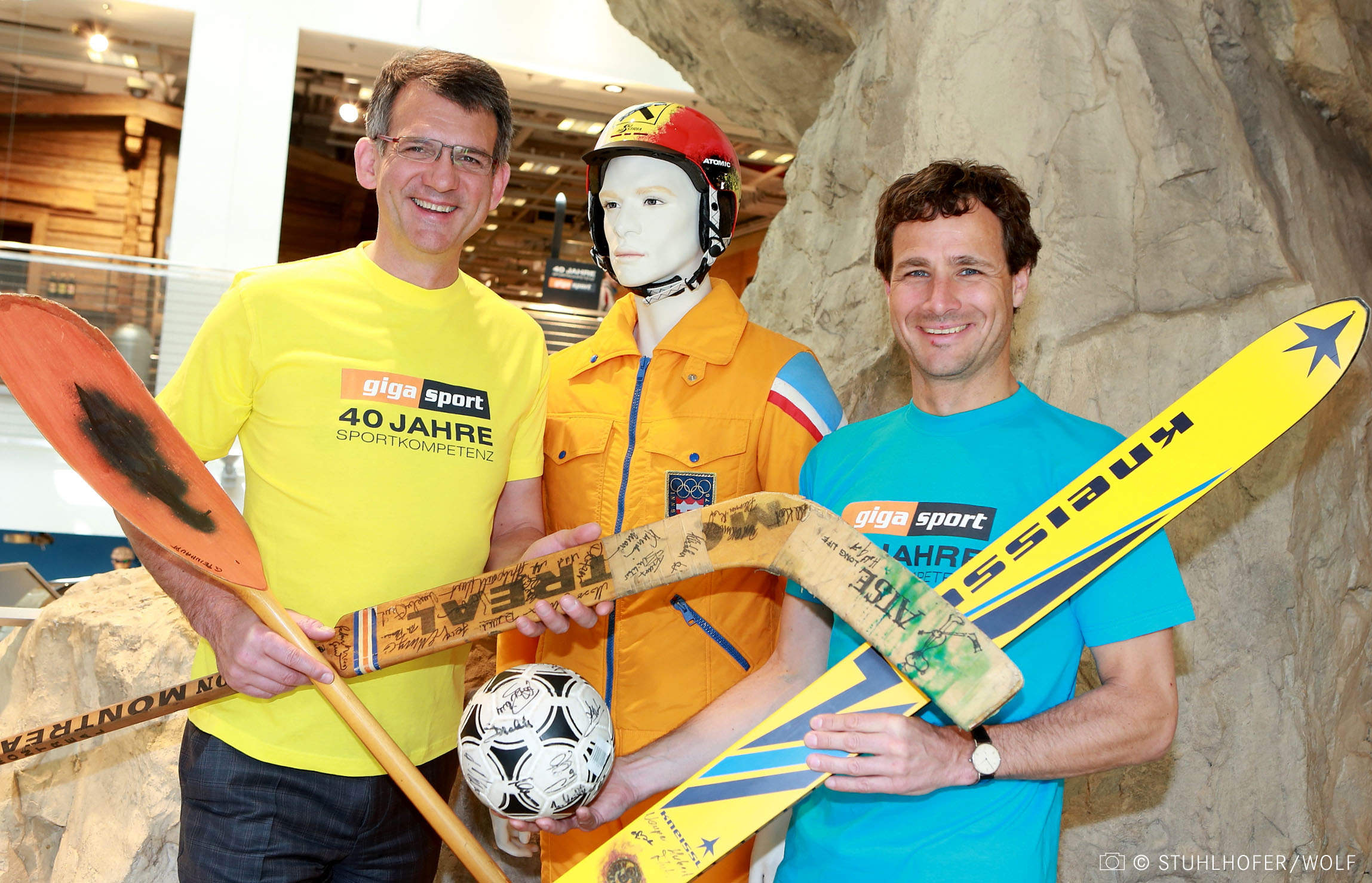 Zwei lächelnde Männer mit Sportgeräten – einem Kajakpaddel und einem Hockeyschläger – stehen neben einer Schaufensterpuppe mit orangefarbener Weste und Helm und einem Fußball in der Hand. Ein Mann trägt ein gelbes Hemd, der andere ein blaues. Im Hintergrund ist ein felsiger Hintergrund zu sehen.