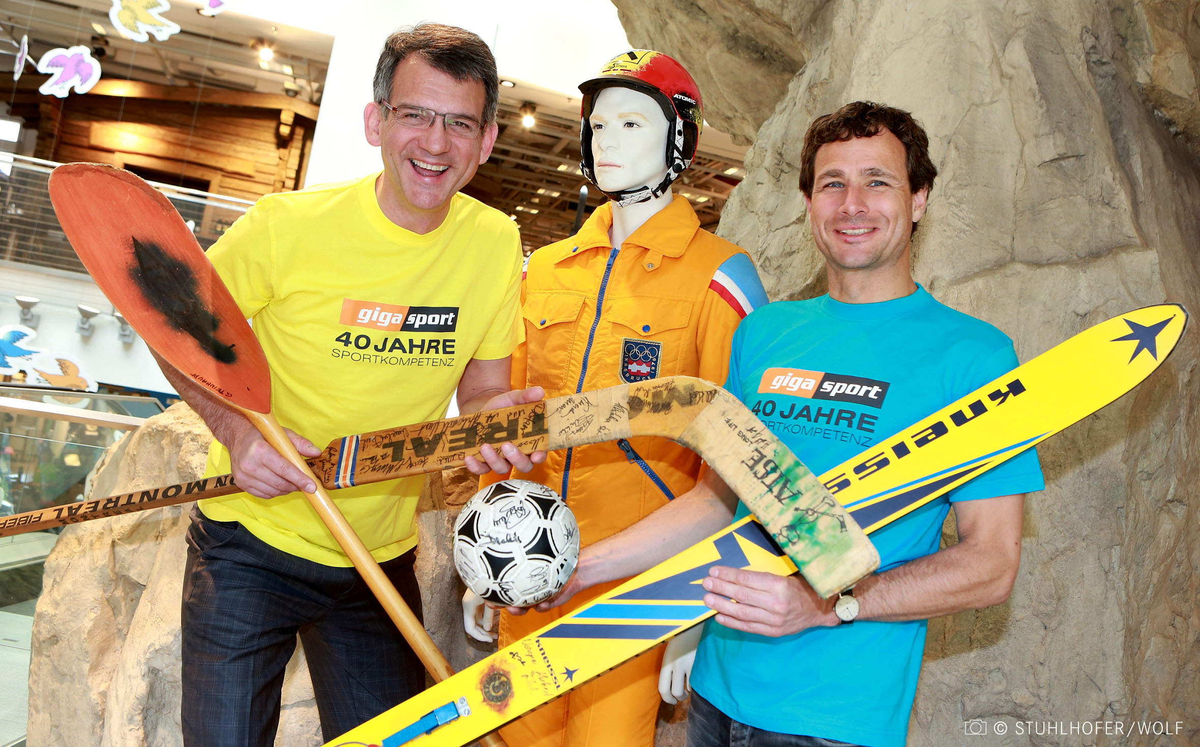 Zwei Männer in hellen Hemden halten Sportgeräte, darunter ein Paddel, Skier und einen Hockeyschläger, neben einer Schaufensterpuppe in gelber Rettungsuniform, die einen Fußball hält. Sie sind drinnen, im Hintergrund ist eine Kletterwand zu sehen.