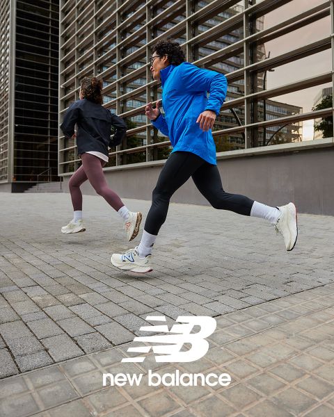 Zwei Personen joggen im Freien, eine in blauer Jacke und schwarzen Leggings, die andere in schwarzer Jacke und kastanienbraunen Leggings. Beide tragen weiße Turnschuhe. Sie laufen neben einem modernen Gebäude mit Glaspaneelen. Unten ist das New Balance-Logo zu sehen.