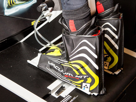 Nahaufnahme eines Paars Skischuhe, das zur individuellen Anpassung in eine Maschine eingespannt ist. An den Schuhen sind Schläuche befestigt, die ein schwarz-gelbes Design mit dem Text „VACUUM FIT“ aufweisen. Die Einrichtung scheint Teil eines speziellen Anpassungsprozesses zu sein.