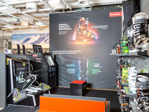 Ausstellung von Skischuhen und Ausrüstung in einem Geschäft. Ein großes Poster zeigt einen orangefarbenen Skischuh mit Text. In den Regalen rechts sind verschiedene Skischuhe ausgestellt. Das Geschäft hat ein helles, modernes Interieur mit Deckenleuchten und weißen Wänden.