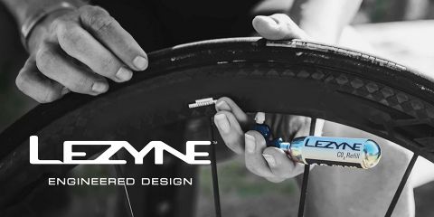 Die Hände einer Person befestigen einen Lezyne CO2-Inflator an einem Fahrradreifen. Der Reifen wird aufgepumpt und auf der Inflatorkartusche ist das Lezyne-Branding zu sehen. Der Text „Lezyne Engineered Design“ ist deutlich im Vordergrund zu sehen.