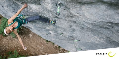 Ein Kletterer hängt an einer steilen Felswand, gesichert mit Kletterausrüstung. Der Kletterer trägt ein grünes Oberteil und eine graue Hose. Der Hintergrund zeigt eine felsige Oberfläche und etwas Grün. Unten rechts ist das Edelrid-Logo zu sehen.