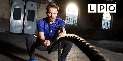 Ein Mann in blauem T-Shirt und dunkler Hose macht in einem Fitnessstudio im Industriestil mit zwei großen Bogenfenstern im Hintergrund eine Battle Rope-Übung. Licht fällt herein und in der oberen rechten Ecke erscheinen die Buchstaben „LPO“.