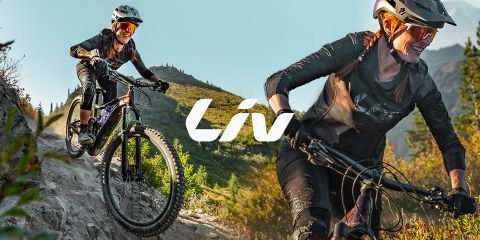 Zwei Frauen fahren mit dem Mountainbike auf einem schroffen Pfad mit Bäumen und Bergen im Hintergrund. Beide tragen Helme und Sonnenbrillen. In der Mitte ist deutlich das Wort „Liv“ zu sehen.