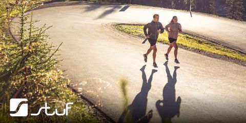 Zwei Menschen joggen auf einer kurvenreichen Straße, die von einem Wald umgeben ist. Die Sonne wirft lange Schatten auf den Bürgersteig. Beide tragen Sportkleidung. In der unteren linken Ecke befindet sich ein Logo mit der Aufschrift „Stuf“.