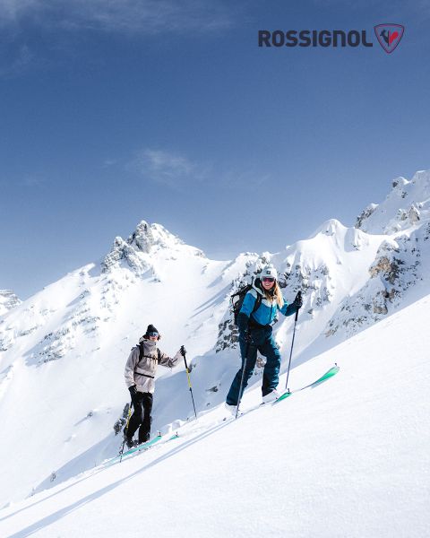 Zwei Skifahrer in Winterkleidung fahren an einem klaren Tag einen verschneiten Berghang hinauf. Im Hintergrund sind schroffe, schneebedeckte Gipfel vor blauem Himmel zu sehen. Das Rossignol-Logo ist in der oberen rechten Ecke zu sehen.