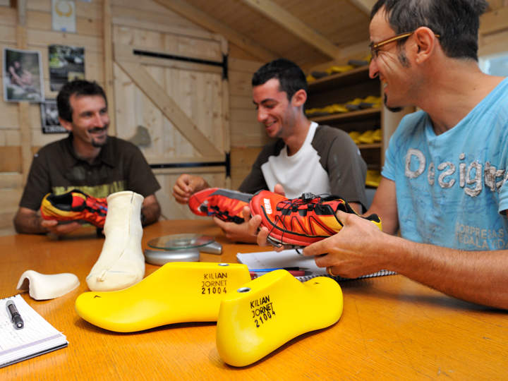 Drei Männer sitzen um einen Tisch herum und halten jeweils unterschiedliche Laufschuhe in den Händen. Auf dem Tisch liegen gelbe Schuhformen mit der Aufschrift „Kilian Jornet“. Im Hintergrund sind Regale mit gelben Formen zu sehen. Sie wirken vertieft und lächeln.