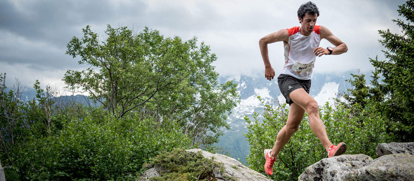 Eine Person in roten Laufschuhen und weiß-rotem Sportoutfit läuft über felsiges Gelände in einem Bergwald. Im Hintergrund sind bewölkter Himmel und schneebedeckte Gipfel zu sehen.