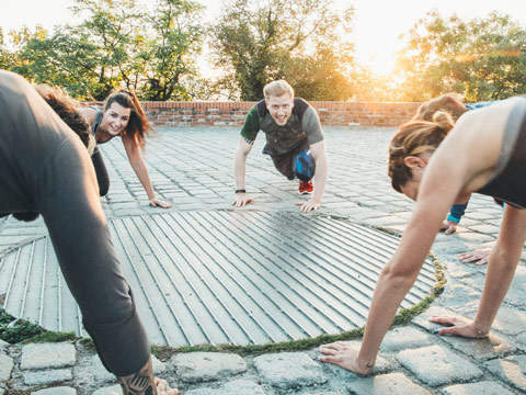 Eine Gruppe von Menschen macht im Freien auf einer Kopfsteinpflasterfläche um ein rundes Metallgitter herum Liegestütze. Sie lächeln und im Hintergrund geht die Sonne unter und wirft einen warmen Schein. Bäume und eine Backsteinmauer sind zu sehen.