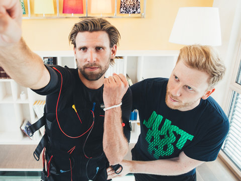Zwei Männer stehen in einem Raum mit Regalen und bunten Büchern im Hintergrund. Ein Mann trägt einen schwarzen Anzug mit elektronischen Sensoren und zeigt nach vorne, während der andere Mann in einem dunklen T-Shirt neben ihm steht und seinen Arm hält.