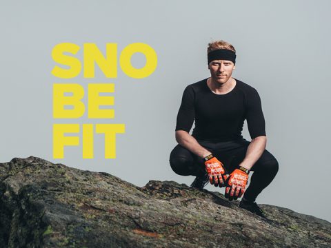 Eine Person in schwarzem Outfit und Stirnband hockt draußen auf Felsen. Neben ihr steht der gelbe Text „SNO BE FIT“. Sie trägt orangefarbene Handschuhe und der Hintergrund ist ein grauer Himmel.
