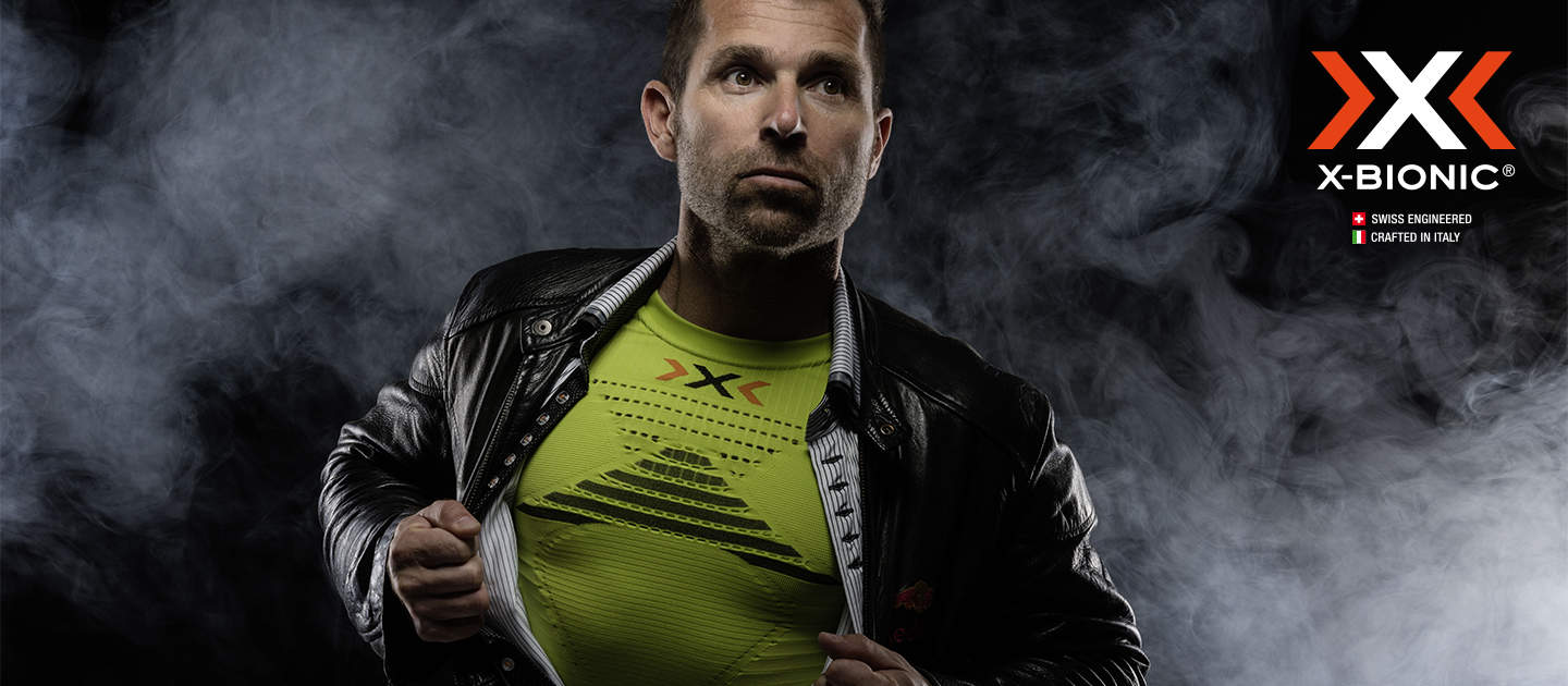 Ein Mann posiert selbstbewusst und öffnet eine schwarze Lederjacke. Darunter ist ein leuchtend grünes X-Bionic-Shirt zu sehen. Im Hintergrund sind Raucheffekte zu sehen, rechts ist das X-Bionic-Logo zu sehen.
