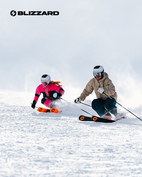Zwei Skifahrer in Winterkleidung fahren einen schneebedeckten Hang hinunter. Der linke Skifahrer trägt ein rosa Outfit, der rechte ein beiges. Beide tragen Helme und Schutzbrillen. Das Blizzard-Logo ist oben links zu sehen.