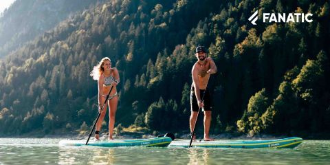Eine Frau im Bikini und ein Mann in Shorts und Hut paddeln auf einem ruhigen See, der von bewaldeten Bergen umgeben ist. Sie sehen beide glücklich und entspannt aus. In der oberen rechten Ecke ist das Logo „FANATIC“ zu sehen.
