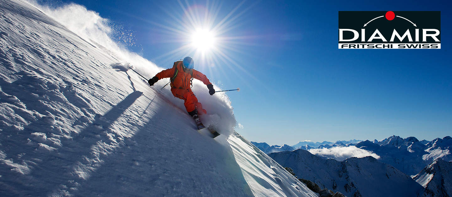Ein Skifahrer in orangefarbenem Outfit fährt durch den Neuschnee auf einem sonnigen, schneebedeckten Berghang. Die Sonne scheint hell am blauen Himmel und erleuchtet die umliegenden schneebedeckten Gipfel. In der oberen rechten Ecke befindet sich ein Logo von „Diamir Fritschi Swiss“.