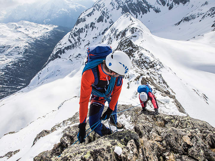 Zwei Bergsteiger erklimmen einen schneebedeckten Bergkamm. Der Vorsteiger trägt eine rote Jacke, blaue Hosen und einen weißen Helm und sichert sich mit Seilen. Der zweite Bergsteiger folgt ihm dicht auf den Fersen. Schneebedeckte Gipfel und Täler erstrecken sich unter einem klaren Himmel.