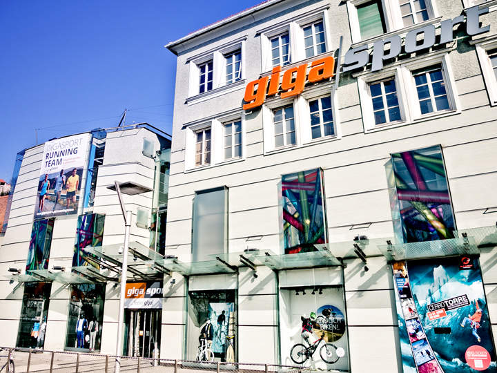 Straßenansicht des Gigasport-Geschäfts mit großen Glasfenstern, in denen Sportprodukte ausgestellt sind. Auf einer Werbetafel sind Läufer abgebildet. Das Gebäude hat eine moderne Fassade mit „Gigasport“ in kräftigen orangefarbenen und grauen Buchstaben.