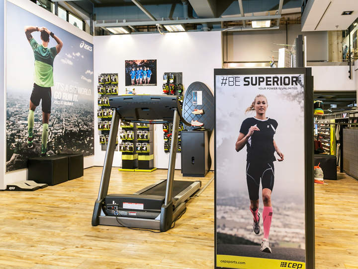 Ladeneinrichtung mit Holzboden, Ausstellung von Sportartikeln und einem Laufband. Poster an den Wänden und ein Standschild zeigen Sportler in Sportbekleidung. Helles, modernes Ambiente mit verschiedenen Fitness-Accessoires in den Regalen.