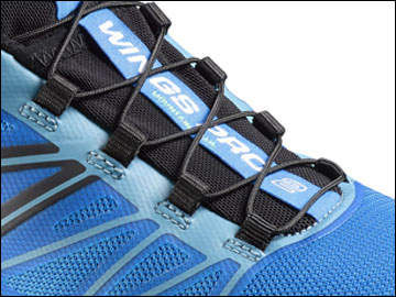 Nahaufnahme eines blauen Sportschuhs mit schwarzen Schnürsenkeln und schwarz-blauen Designdetails. Das Schnürsystem besteht aus überkreuzten Riemen und einer kleinen Schlaufe mit der Aufschrift „WINGS“. Das Schuhmaterial wirkt strukturiert und leicht.